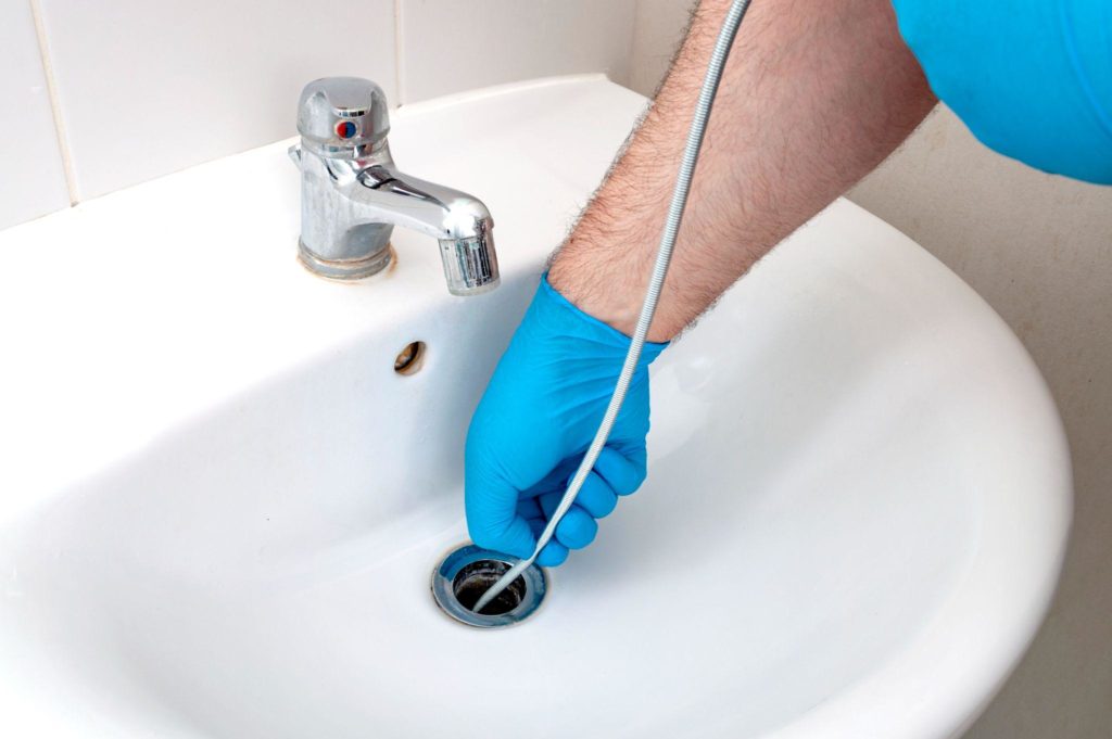 Plumber wearing blue gloves uses a tool to clear a bathroom sink drain