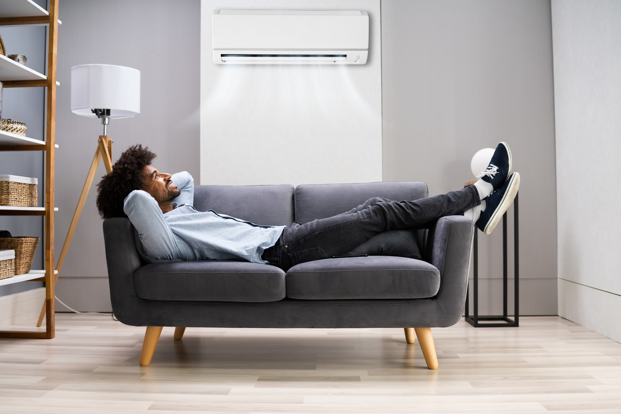 A young Black man relaxing on a couch under an air conditioner blowing air.